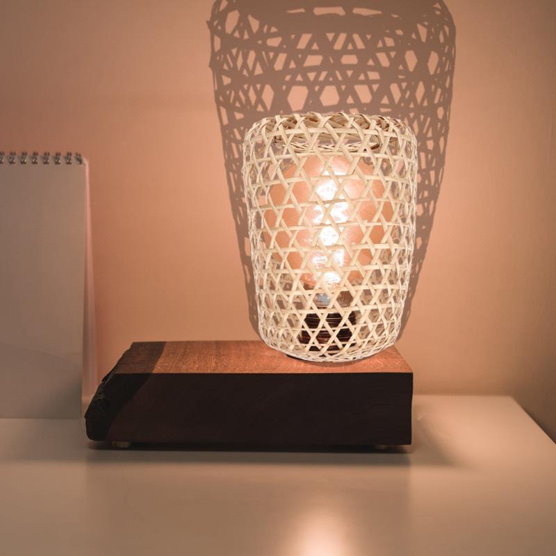 Bloc de bois avec une grosse ampoule allumée et un petit abat-jour en rotin tressé aéré. posé sur une table blanche. On voit sur la gauche un bloc note posé contre le mur qui est rose. Et on voit les ombres de l'abat-jour sur le mur derrière la lampe.