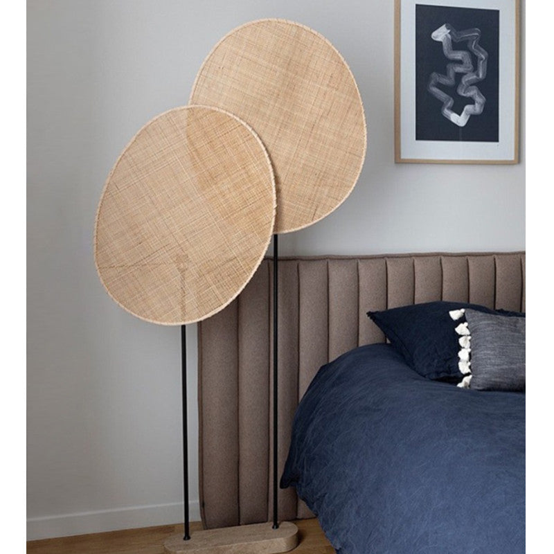 Lampadaire LED autoporté en rotin en forme de grand pétale de fleur, monté sur 2 pieds fins en fer chacun d'une hauteur différentes sur socle bois rectangle arrondit. Mise en scène dans un angle de chambre, ou l'on voit une tête de lit marron, un bout de lit drap et coussin bleu et un cadre accroché au mur.