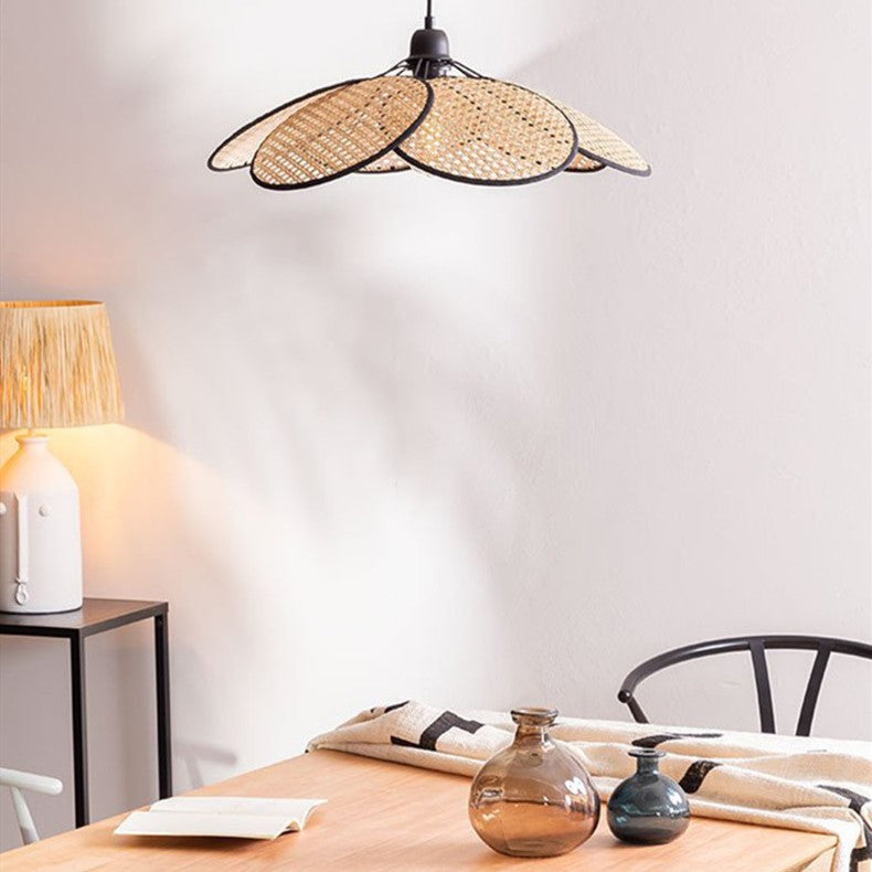 Pièce blanche, avec un luminaire abat-jour en rotin qui se compose de plusieurs pales ronde finition anneau noir, ouvert tel une fleur. En dessous se trouve une table rectangle en bois, avec deux vase boules en verre marron et noir, un petit livre ouvert et un tissus blanc avec des motifs noir poussé en bord de table, on voit un bout de dossier de chaise en métal et sur la gauche de l'image on voit un bout de desserte en métal noir, avec une petite lampe allumée blanche avec un abat-jour beige.