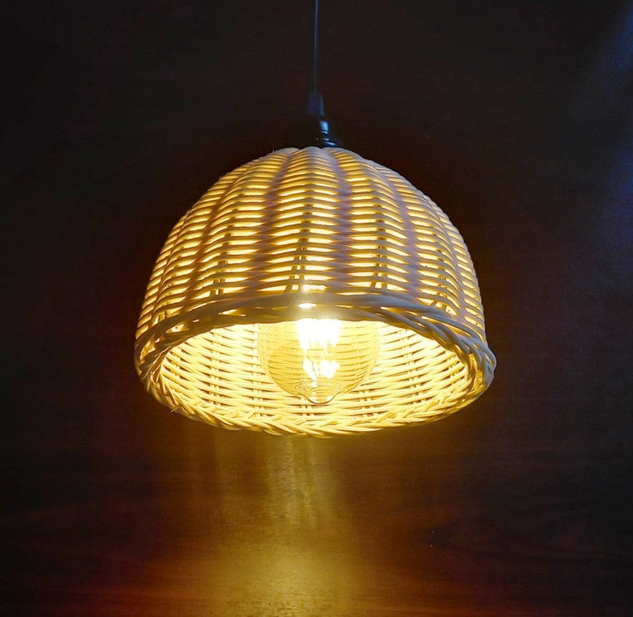 Abat-jour en rotin épais tressé en forme de panier à l'envers, devant un mur en bois foncé dans le noir. On voit une grosse ampoule allumé dans l'abat-jour, et un peu du fil qui le suspend.