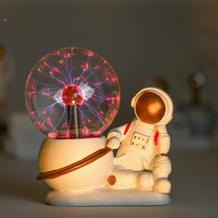 Socle blanc représentant un astronaute assit, une jambe pliée horizontalement et collée au sol, l'autre jambe pliée verticalement avec le pied au sol et son coude posé sur son genoux car son bras et lui aussi plié et tien sa tête. L'autre bras et accoudé sur une planète blanche avec un anneau doré, sur laquelle est présenté la lampe boule plasma allumée avec plusieurs faisceaux bleu et rouge. 