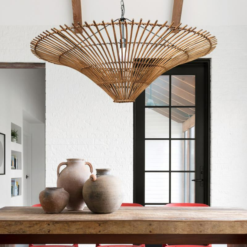 Photo avec grande table rectangle en bois massif, avec 3 jarres de taille différente en terre cuite. Au dessus un grand lustre artisanal en tige de rotin en forme de cône, serré en bas et très large en haut. derrière un mur blanc avec une porte fenêtre sur la droite et une ouverture sue la gauche, on y voit trois trous carré dans le mur côté gauche avec de la décoration 